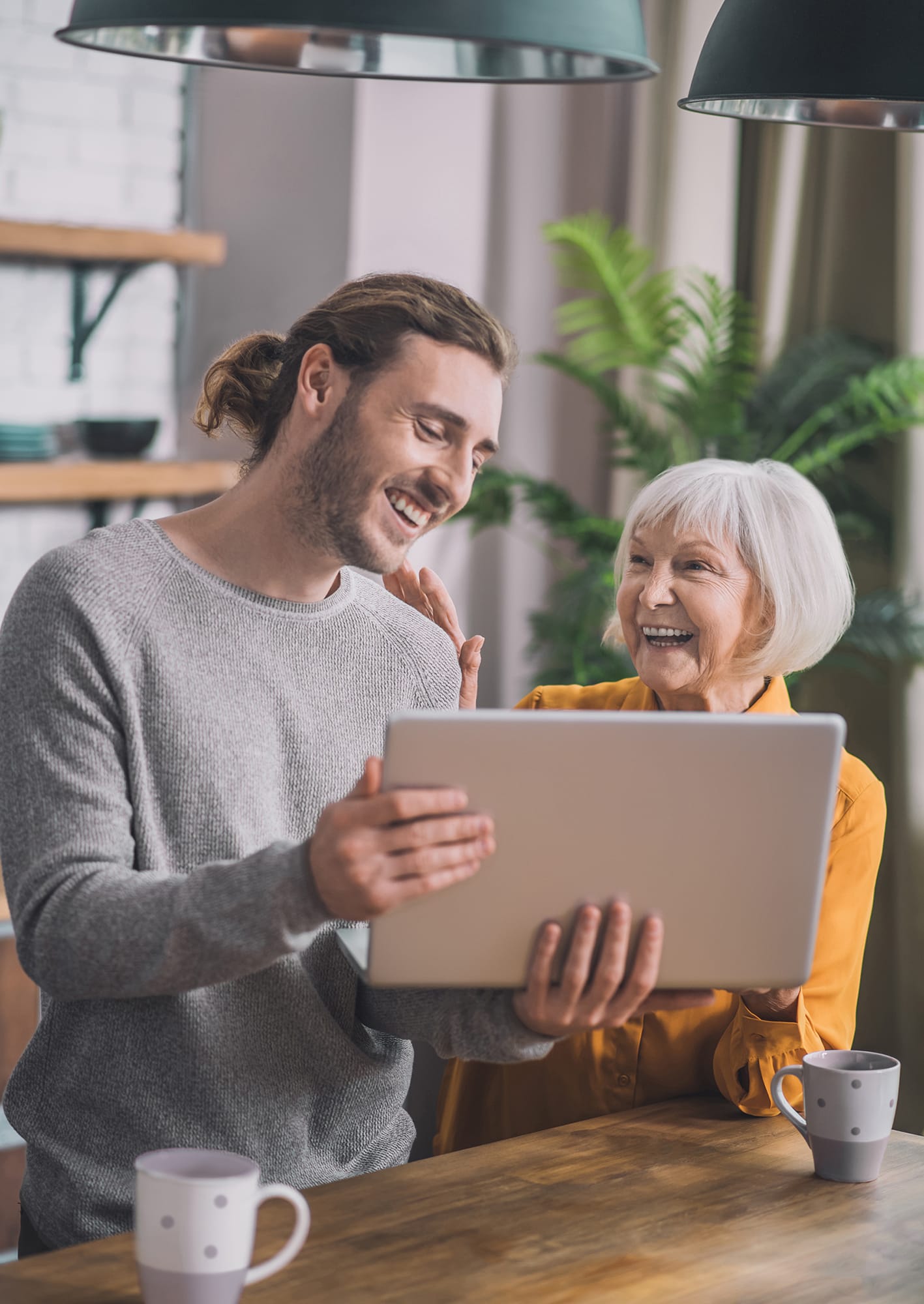 makeup-studio-33 Smiling young man showing an older woman something on a laptop while standing together in a cozy kitchen.